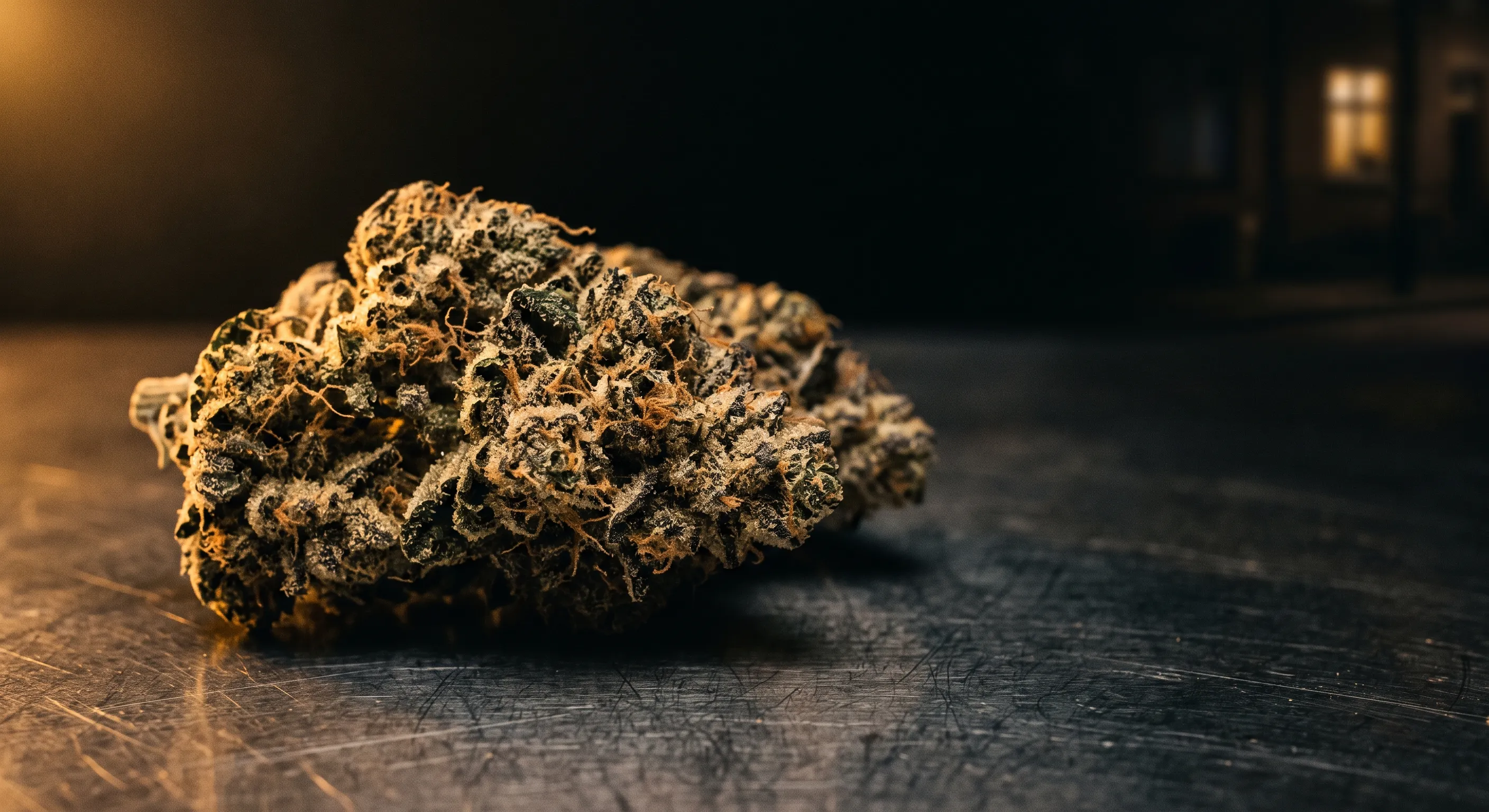 Cannabis flower on a steel counter under warm light, with a lit window in the darkness behind